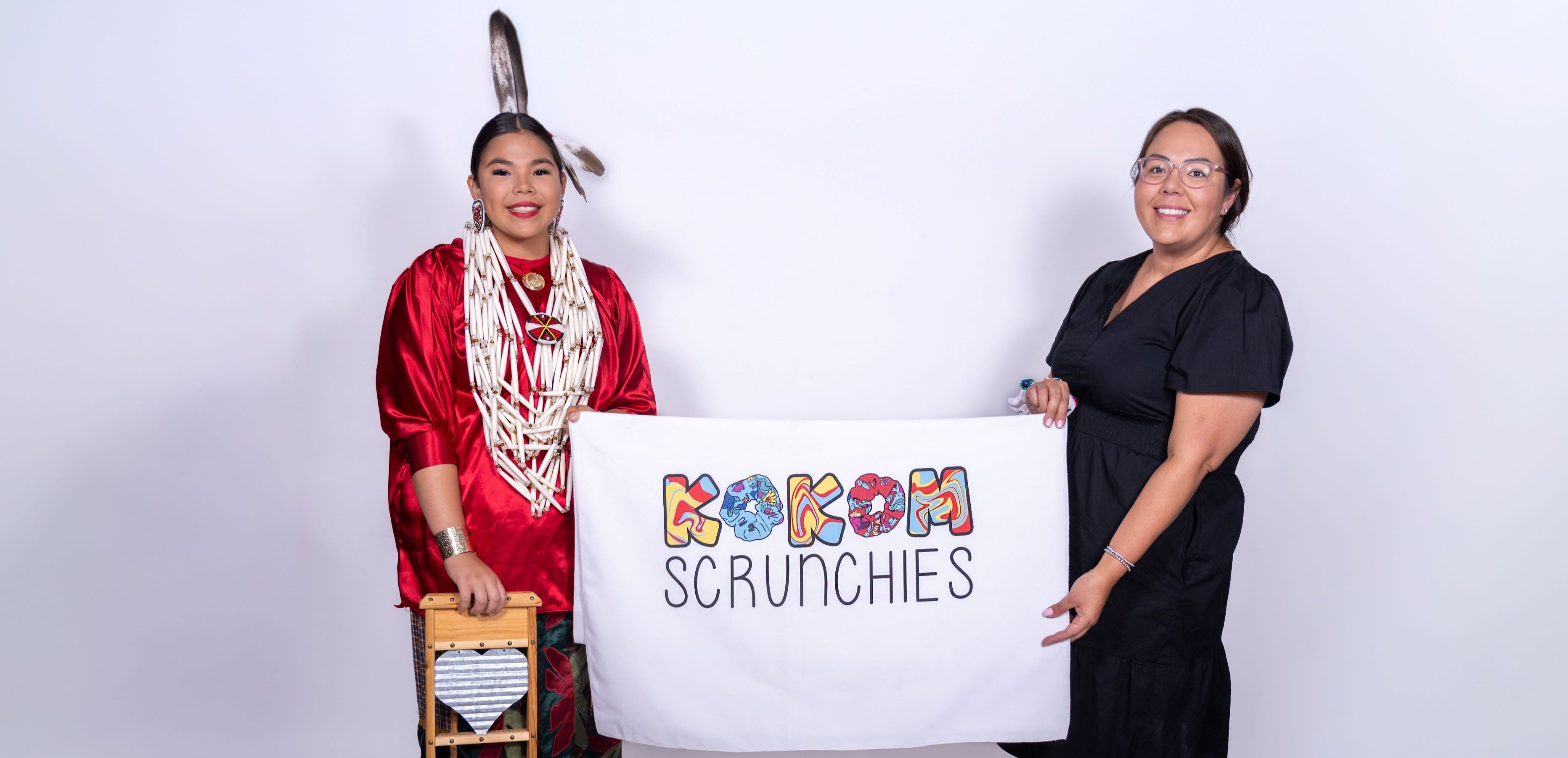 Two women holding a sign with 'kokom Scrunchies' on a white background