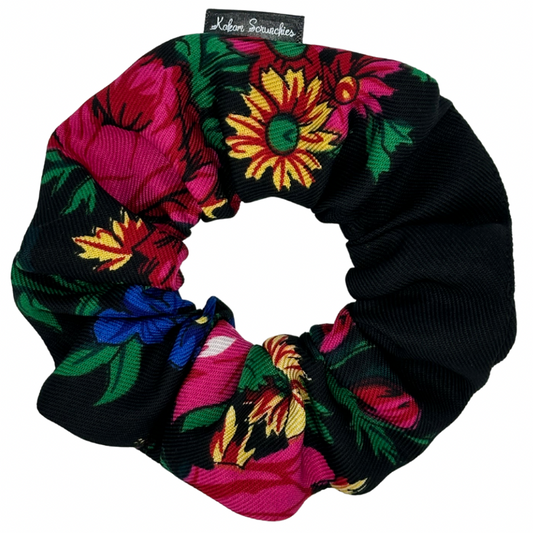 A black scrunchie with a vibrant floral pattern consisting of large flowers in red, pink, yellow, and blue on a tag, against a white background.