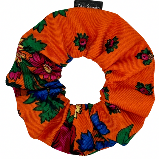 An orange hair scrunchie with a floral pattern, including green leaves and purple flowers.