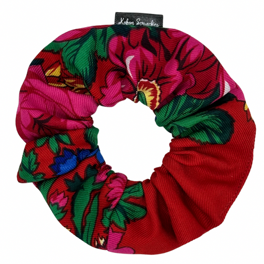 A red scrunchie with a floral pattern, featuring tags with brand name 'Kokom Scrunchies' visible.