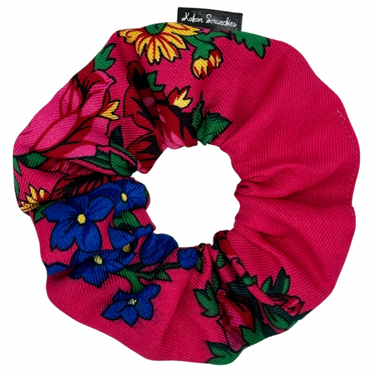 A pink hair scrunchie with a floral pattern, including blue and yellow flowers, and green leaves.